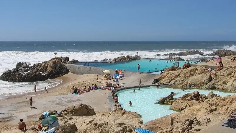 A sunny coastal beach with natural tidal pools nestled among rocky cliffs, where people are swimming, relaxing, and enjoying the ocean views as waves crash against the shoreline.