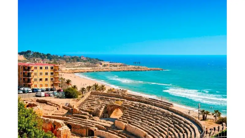 Ancient Roman amphitheater overlooking a sandy beach with turquoise Mediterranean waters under a clear blue sky.