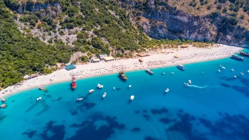 A stunning aerial view of a white-sand beach with crystal-clear turquoise water, anchored boats, and a lush green hillside creating a picturesque Mediterranean coastal landscape.