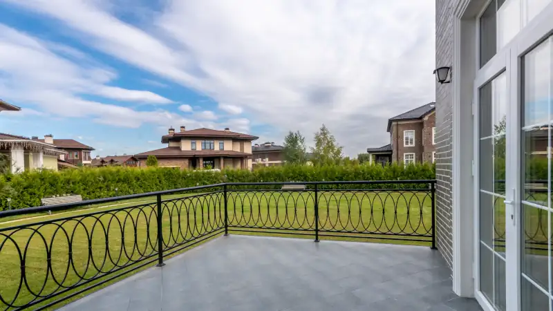 The scene shows a balcony with a view of multiple houses and a large green lawn. The sky has clouds and is bright with sunlight. It is a peaceful day in the suburbs