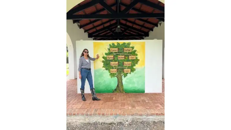 A woman stands beside a colorful mural featuring a large tree with wooden-style signposts displaying names