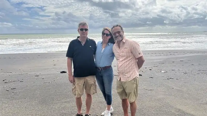Lief, Kathleen and a man posing together on a sandy beach with ocean waves and dramatic clouds in the background.