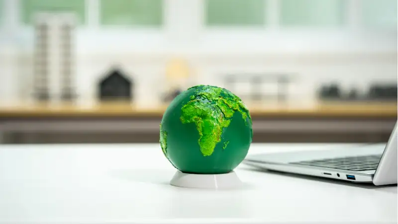 Green globe on a desk beside a laptop with a blurred home office background