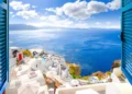 Hillside view through an open window with blue shutters of the caldera, sea and white village of Oia on the island of Santorini, Greece.