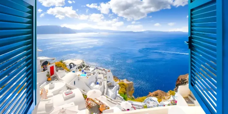 Hillside view through an open window with blue shutters of the caldera, sea and white village of Oia on the island of Santorini, Greece.