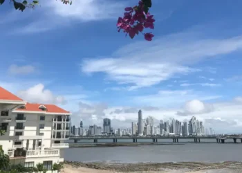 A scenic waterfront view of Panama City’s modern skyline, with a red-roofed building and pink bougainvillea framing the bay beneath a bright blue sky.
