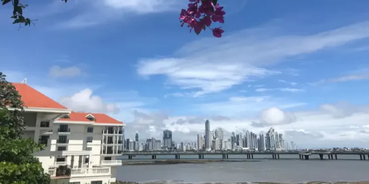 A scenic waterfront view of Panama City’s modern skyline, with a red-roofed building and pink bougainvillea framing the bay beneath a bright blue sky.