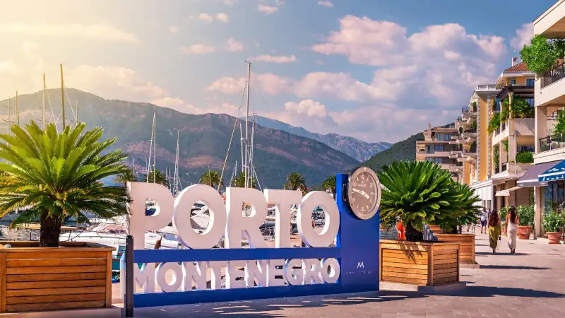 Porto Montenegro luxury marina village entrance featuring the iconic sign, promenade, palm trees, and Mediterranean architecture.