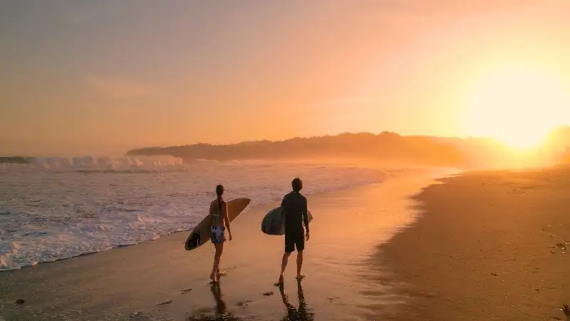Surfers walking along a beach at sunset, carrying their boards as golden light reflects on the wet sand.