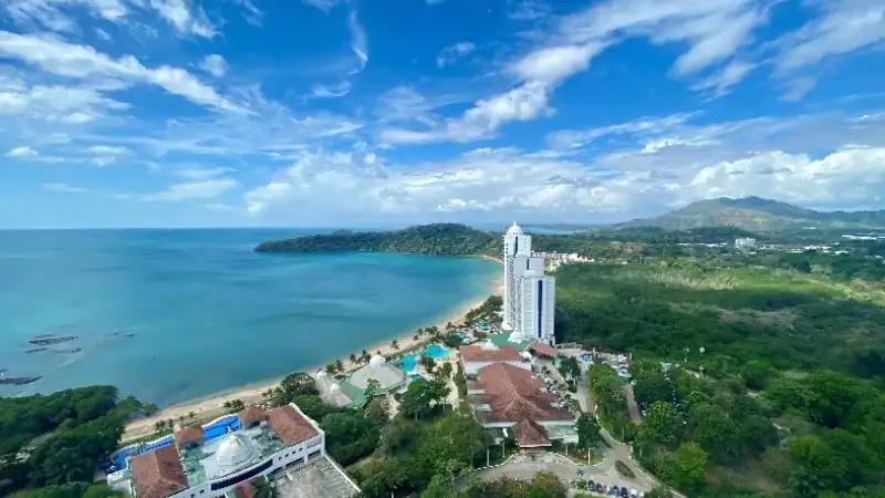 Aerial view of a tropical coastal resort overlooking a beautiful turquoise bay, surrounded by lush greenery and mountains.