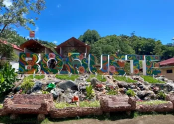 Colorful “Boquete” sign surrounded by lush greenery, tropical flowers, and rustic seating under a bright blue sky.