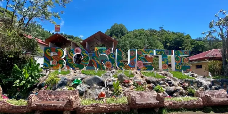Colorful “Boquete” sign surrounded by lush greenery, tropical flowers, and rustic seating under a bright blue sky.