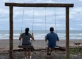 Kathleen and Lief relaxing on a wooden swing overlooking a quiet beach under a cloudy sky.