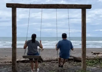 Kathleen and Lief relaxing on a wooden swing overlooking a quiet beach under a cloudy sky.