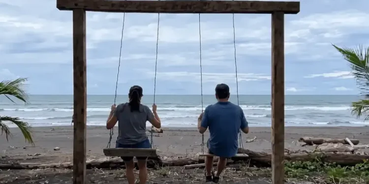 Kathleen and Lief relaxing on a wooden swing overlooking a quiet beach under a cloudy sky.