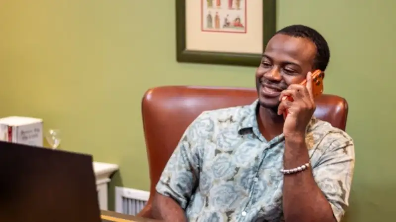 Justin smiles while talking on his phone, seated at a desk with a laptop in a warmly lit office.