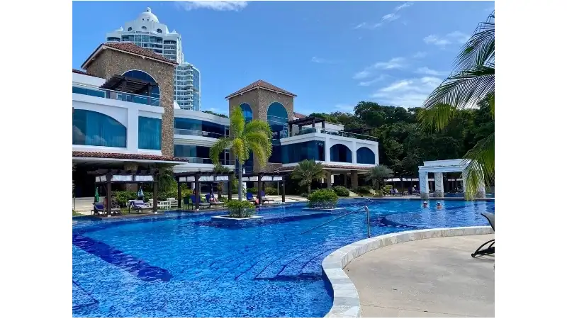 A luxurious resort swimming pool featuring elegant architecture, palm trees, and modern outdoor lounging areas under a clear blue sky.