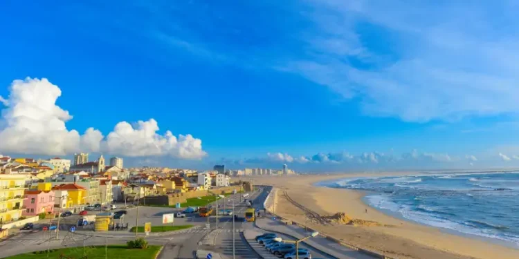 Sunny coastal cityscape in Portugal with colorful buildings, a sandy beach, rolling Atlantic waves, and a vivid blue sky.