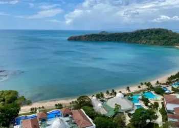 Stunning aerial view of a tropical beach resort featuring turquoise waters, lush green coastline, white sandy beaches, and a bright blue sky dotted with clouds.