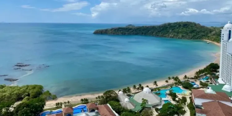 Stunning aerial view of a tropical beach resort featuring turquoise waters, lush green coastline, white sandy beaches, and a bright blue sky dotted with clouds.