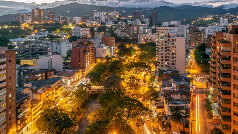 Cityscape of Cali, Colombia at dusk, with lit streets, tall buildings, and mountains in the background.