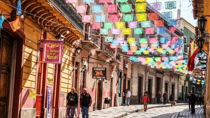 Mexicans walking on cobblestone street with sun lit colorful banner flags hanging above, Guanajuato